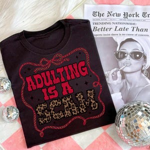 May include: Black t-shirt featuring the slogan "ADULTING IS A SCAM" in red and leopard print. The design is enclosed in a decorative red border. The shirt is placed on a pink and white checkered surface.