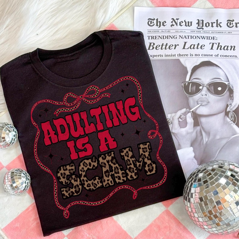 May include: Black t-shirt featuring the slogan "ADULTING IS A SCAM" in red and leopard print. The design is enclosed in a decorative red border. The shirt is placed on a pink and white checkered surface.