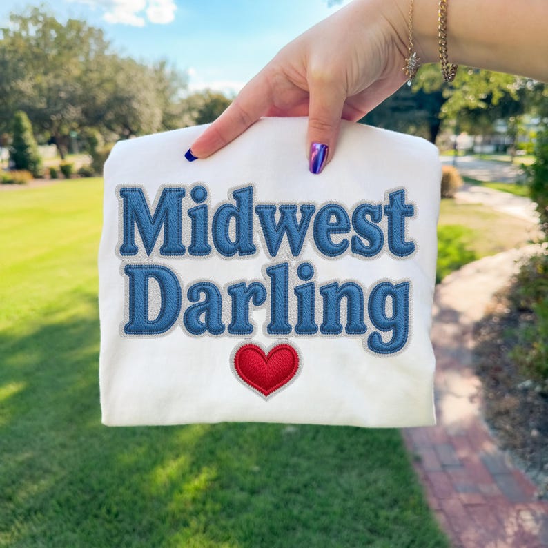 May include: White sweatshirt with the embroidered text "Midwest Darling" in blue denim-style letters. A red heart is embroidered below the text. The sweatshirt is held up against a background of green grass and trees.