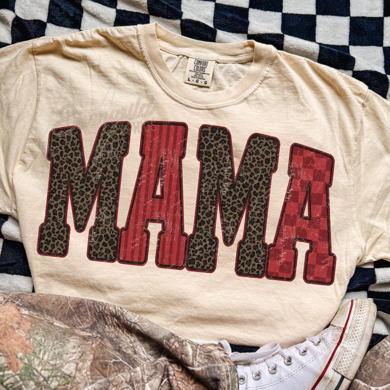 May include: A cream-colored short-sleeve t-shirt featuring the word MAMA in large block letters. The letters M and M have a brown leopard print, while the letters A and A feature red vertical stripes and a red checkered pattern respectively.