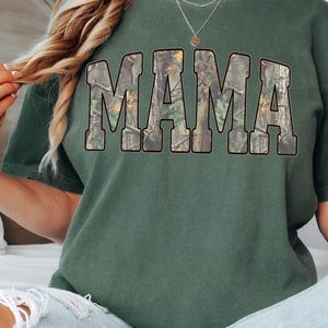 May include: Green short-sleeved t-shirt with the word "MAMA" in large, block letters across the front. The letters are filled with a camouflage pattern. The shirt is a solid green color.