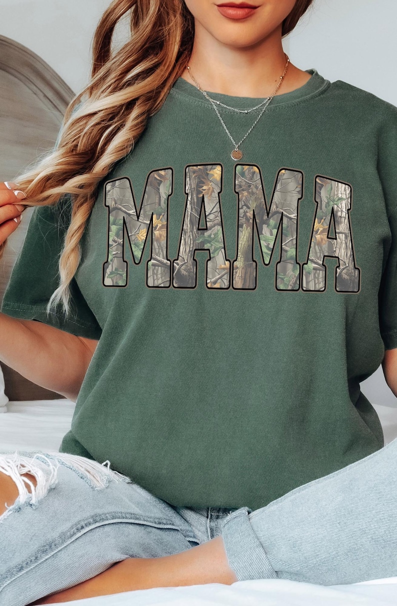 May include: Green short-sleeved t-shirt with the word "MAMA" in large, block letters across the front. The letters are filled with a camouflage pattern. The shirt is a solid green color.