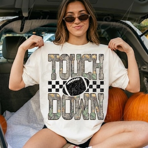 May include: A cream-colored short-sleeve t-shirt featuring the text TOUCH DOWN in a camouflage pattern. A black and white checkered stripe separates the words, with a black football graphic centered between them. The design has a distressed texture.