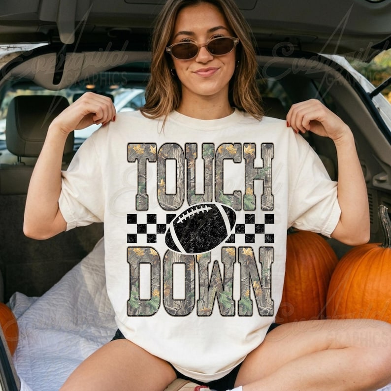 May include: A cream-colored short-sleeve t-shirt featuring the text TOUCH DOWN in a camouflage pattern. A black and white checkered stripe separates the words, with a black football graphic centered between them. The design has a distressed texture.