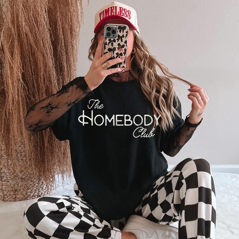 K&ouml;nnte beinhalten: Schwarzes T-Shirt mit dem wei&szlig;en Schriftzug "The HOMEBODY Club". Die Person tr&auml;gt eine Baseballkappe mit "TIMELESS" und eine schwarz-wei&szlig; karierte Hose. Die &Auml;rmel sind aus durchsichtiger schwarzer Spitze.