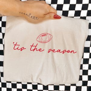 May include: A cream-colored t-shirt featuring a red line-art illustration of an American football centered above the red cursive text that reads 'tis the season'. The design is printed on the front of the fabric, which is laid flat.