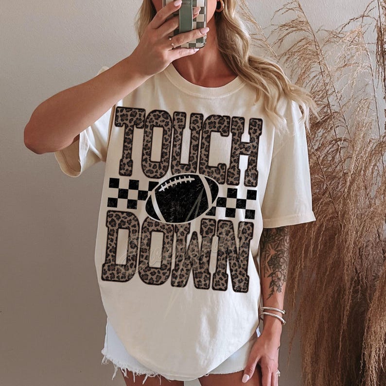 Puede incluir: Una camiseta de manga corta color crema con el texto TOUCH DOWN en un estampado de leopardo marr&oacute;n. Entre las palabras hay un gr&aacute;fico de un bal&oacute;n de f&uacute;tbol americano en blanco y negro con un patr&oacute;n de cuadros blanco y negro.