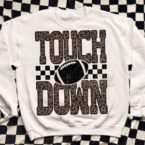 Puede incluir: Una sudadera blanca de cuello redondo con un estampado gr&aacute;fico que incluye la palabra TOUCH en un patr&oacute;n de leopardo marr&oacute;n y negro, una ilustraci&oacute;n de un bal&oacute;n de f&uacute;tbol americano negro y la palabra DOWN en estampado de leopardo a juego.