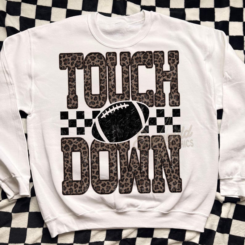 Puede incluir: Una sudadera blanca de cuello redondo con un estampado gr&aacute;fico que incluye la palabra TOUCH en un patr&oacute;n de leopardo marr&oacute;n y negro, una ilustraci&oacute;n de un bal&oacute;n de f&uacute;tbol americano negro y la palabra DOWN en estampado de leopardo a juego.