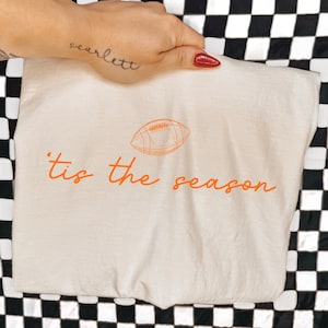 May include: A cream-colored t-shirt featuring an orange graphic of an American football centered above the orange cursive text 'tis the season'. The design is printed on the front of the fabric, which is displayed flat against a black and white checkered background.