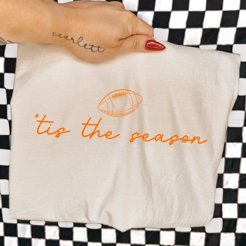 May include: A cream-colored t-shirt featuring an orange graphic of an American football centered above the orange cursive text 'tis the season'. The design is printed on the front of the fabric, which is displayed flat against a black and white checkered background.