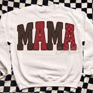 May include: A white crewneck sweatshirt featuring the word MAMA in large block letters. The letters M and M have a brown leopard print, while the A and A feature red vertical stripes and a red checkered pattern with dark outlines.