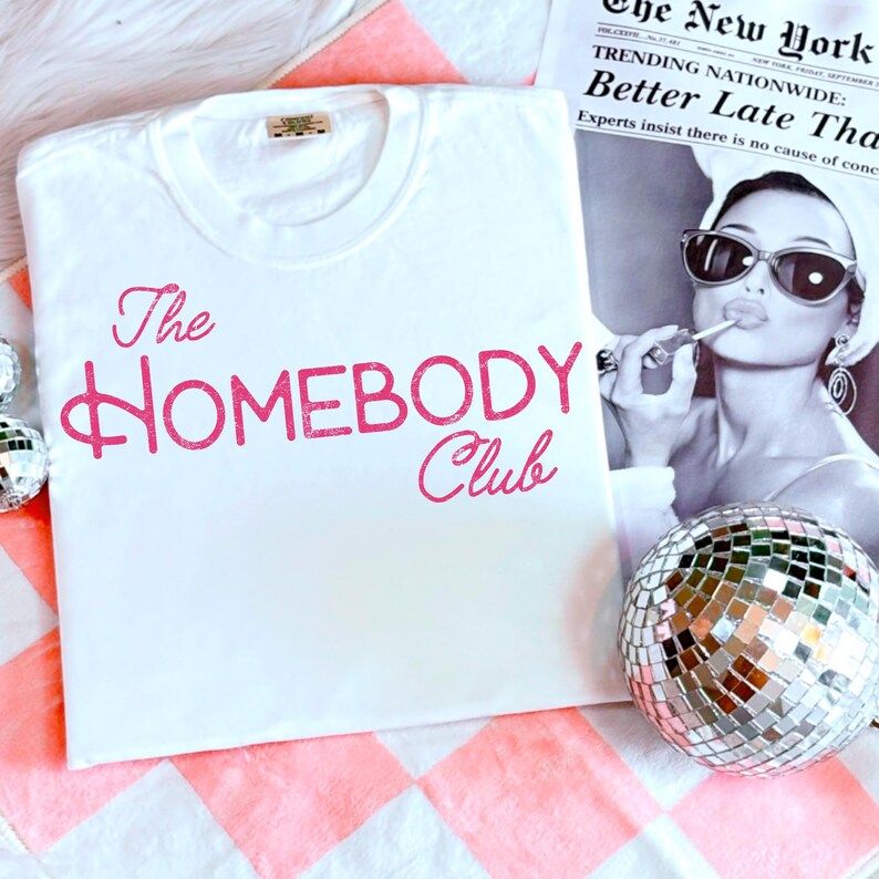 K&ouml;nnte beinhalten: Ein wei&szlig;es T-Shirt mit dem rosa Aufdruck "The Homebody Club". Das T-Shirt liegt auf einer rosa-wei&szlig; karierten Oberfl&auml;che. Eine Discokugel und eine Vintage-Zeitung sind im Hintergrund zu sehen.