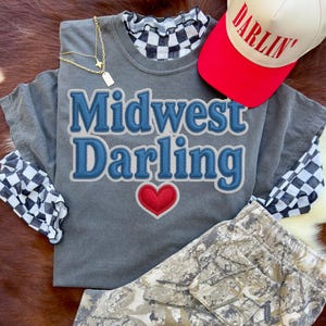 May include: A gray t-shirt with the words "Midwest Darling" in blue and a red heart. A red and white baseball cap with "Darlin'" printed on it. A gold necklace and camouflage pants are also visible.