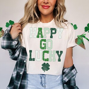 May include: Cream-colored t-shirt with the words "HAPPY GO LUCKY" in green lettering and a four-leaf clover graphic. The model is wearing a green and black plaid shirt over the t-shirt and blue jeans.