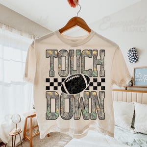 May include: A cream-colored short-sleeve t-shirt featuring the text TOUCH DOWN in a camouflage pattern. A black and white football graphic sits between the words, flanked by a black and white checkered border on a light-colored fabric background.