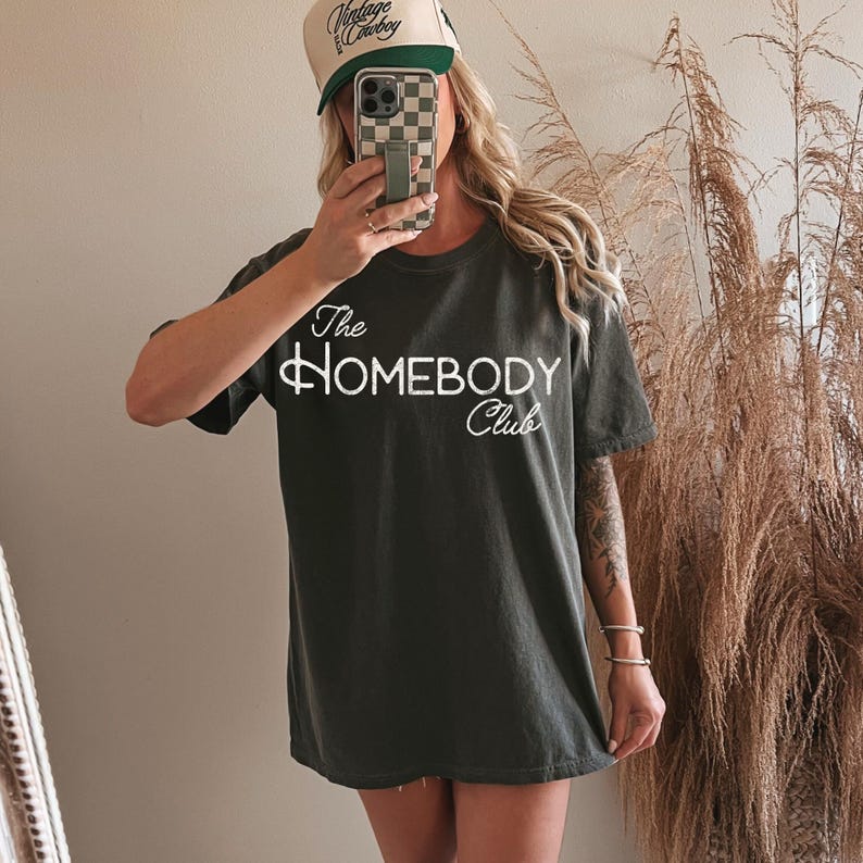 K&ouml;nnte beinhalten: Dunkelgraues T-Shirt mit dem wei&szlig;en Schriftzug "The Homebody Club". Die Person im Bild tr&auml;gt eine beigefarbene Trucker-M&uuml;tze mit den Worten "Vintage Cowboy" in Gr&uuml;n und Schwarz. Die Person h&auml;lt ein Telefon mit kariertem Geh&auml;use.