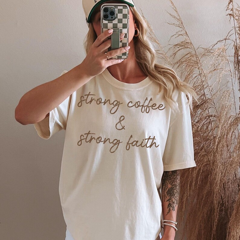 May include: Cream-colored t-shirt with the text "strong coffee & strong faith" in a brown script. The shirt has short sleeves and a relaxed fit. The person in the image is wearing a checkered baseball cap.