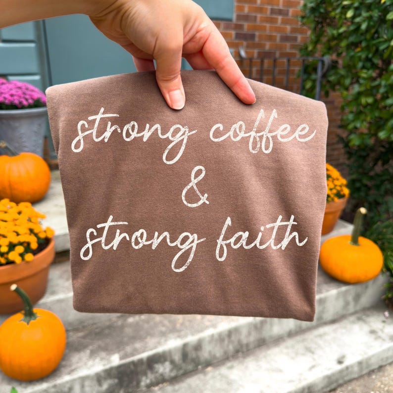 May include: A brown t-shirt with the words "strong coffee & strong faith" in white script. The shirt is held up in front of a background with pumpkins and potted flowers, suggesting a fall theme.