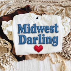 May include: White sweatshirt with the text "Midwest Darling" in blue, stitched lettering, and a red heart. The sweatshirt is folded on top of other clothing items, including a brown and black plaid blanket and a tan sweater.