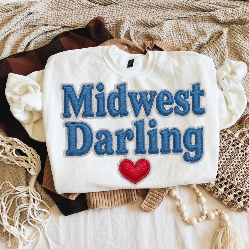 May include: White sweatshirt with the text "Midwest Darling" in blue, stitched lettering, and a red heart. The sweatshirt is folded on top of other clothing items, including a brown and black plaid blanket and a tan sweater.