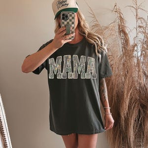 May include: Dark gray t-shirt with the word "MAMA" in a camouflage pattern. The shirt is paired with a trucker hat with the text "Vintage Cowboy" and a green brim. The person is holding a phone with a checkered case.