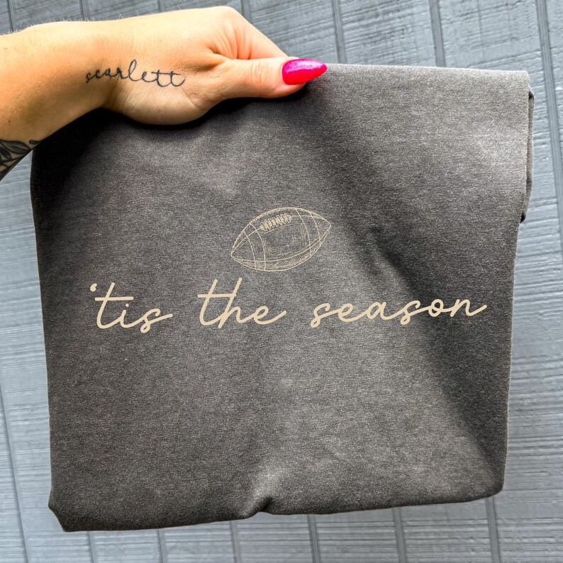 May include: A charcoal grey t-shirt featuring a light tan line-art illustration of an American football above the cursive text 'tis the season' printed in the same light tan color across the center of the garment.