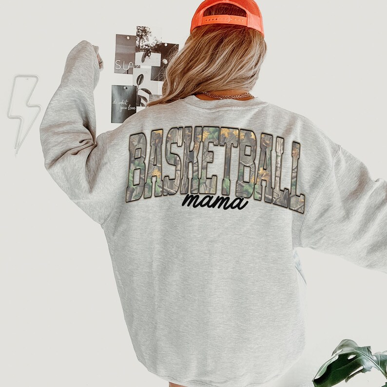 May include: A light grey sweatshirt featuring the text "BASKETBALL mama" in a camouflage pattern. The person in the image is wearing an orange baseball cap.