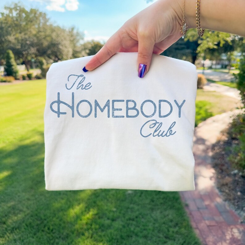 K&ouml;nnte beinhalten: Ein gefaltetes wei&szlig;es T-Shirt mit dem Text "The Homebody Club" in hellblauer Schrift. Das T-Shirt wird vor einem Hintergrund aus gr&uuml;nem Gras und B&auml;umen unter blauem Himmel gehalten.