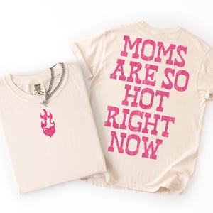 May include: Two cream-colored t-shirts. One shirt has a pink flame heart graphic and a silver chain necklace. The other shirt has the text "MOMS ARE SO HOT RIGHT NOW" printed in pink on the back. The shirts are folded on a white surface.