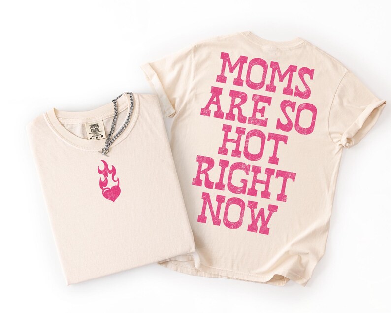 May include: Two cream-colored t-shirts. One shirt has a pink flame heart graphic and a silver chain necklace. The other shirt has the text "MOMS ARE SO HOT RIGHT NOW" printed in pink on the back. The shirts are folded on a white surface.
