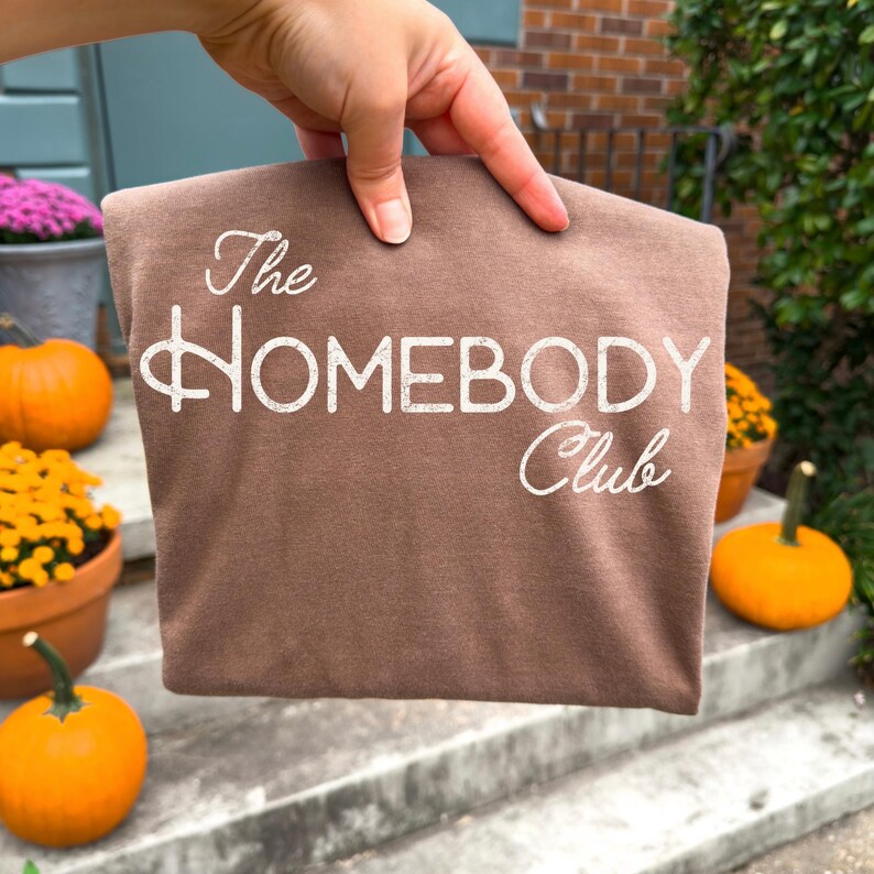 K&ouml;nnte beinhalten: Ein braunes T-Shirt mit dem wei&szlig;en Schriftzug "The Homebody Club" in kursiver Schrift. Das Shirt wird von einer Hand gehalten, mit K&uuml;rbissen und Topfblumen im Hintergrund, was ein Herbstthema suggeriert.