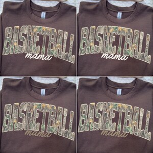 May include: A brown sweatshirt featuring the text "BASKETBALL" in a camouflage pattern and the word "mama" in a cursive font. The sweatshirt is displayed on a wooden hanger.
