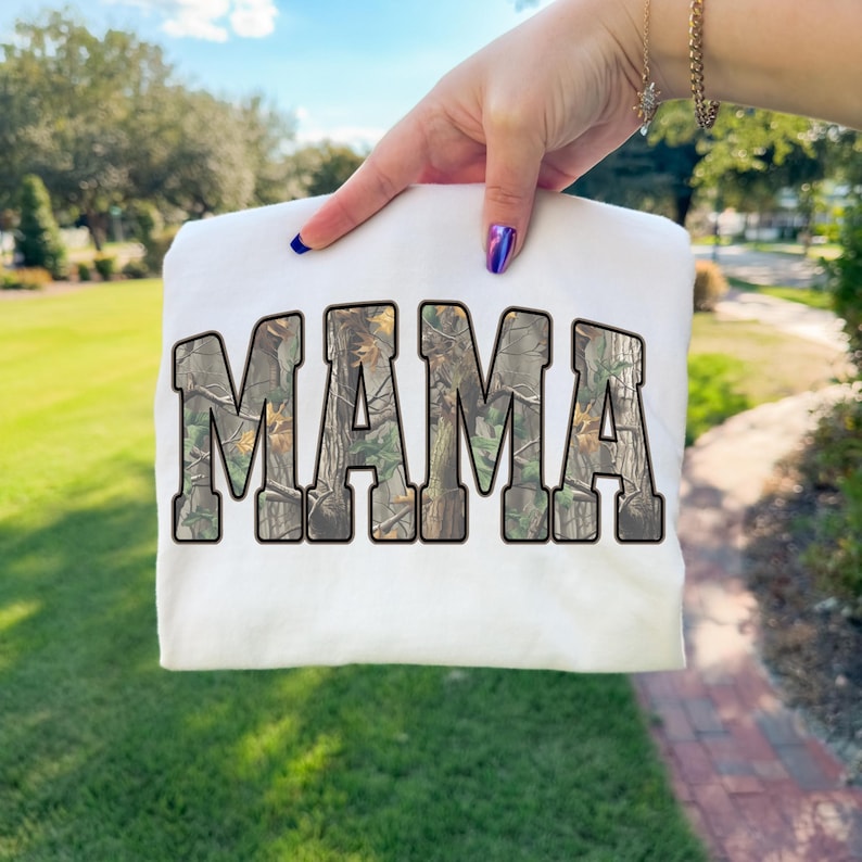 May include: White sweatshirt with the word "MAMA" in large, block letters. The letters are filled with a camouflage pattern in shades of brown, green, and tan. The sweatshirt is held up against a background of green grass and trees.