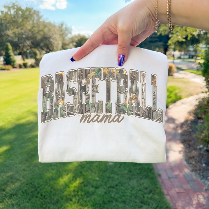 May include: A white sweatshirt featuring the text "BASKETBALL mama" in a camouflage pattern. The garment is held up against a backdrop of green grass and trees, with the sun shining.