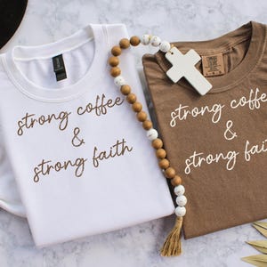 May include: Two t-shirts, one white and one brown, with the phrase "strong coffee & strong faith" printed in a script font. A wooden bead strand with a cross pendant and a tassel is draped between the shirts.