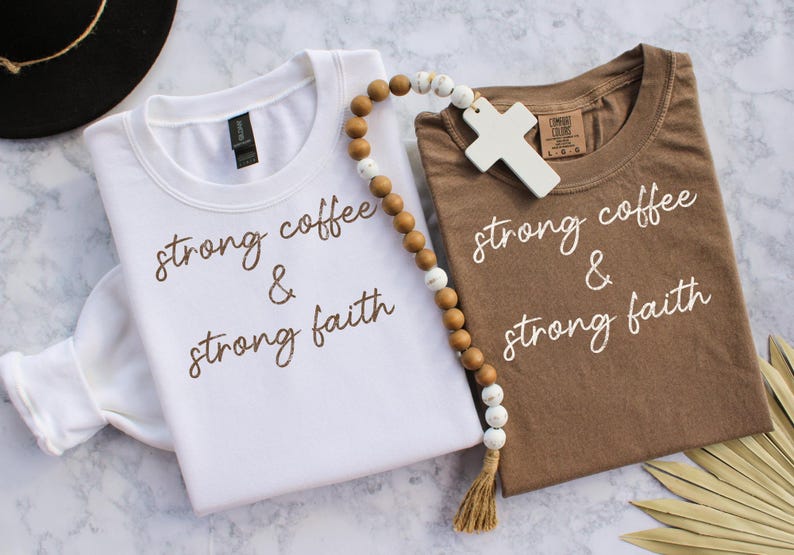 May include: Two t-shirts, one white and one brown, with the phrase "strong coffee & strong faith" printed in a script font. A wooden bead strand with a cross pendant and a tassel is draped between the shirts.