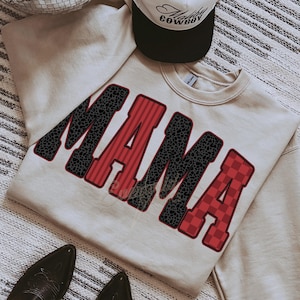 May include: A beige crewneck sweatshirt featuring the word MAMA in large block letters. The letters alternate between a black leopard print pattern and a red striped or checkered pattern, all outlined in a dark red border. A cap is placed above.