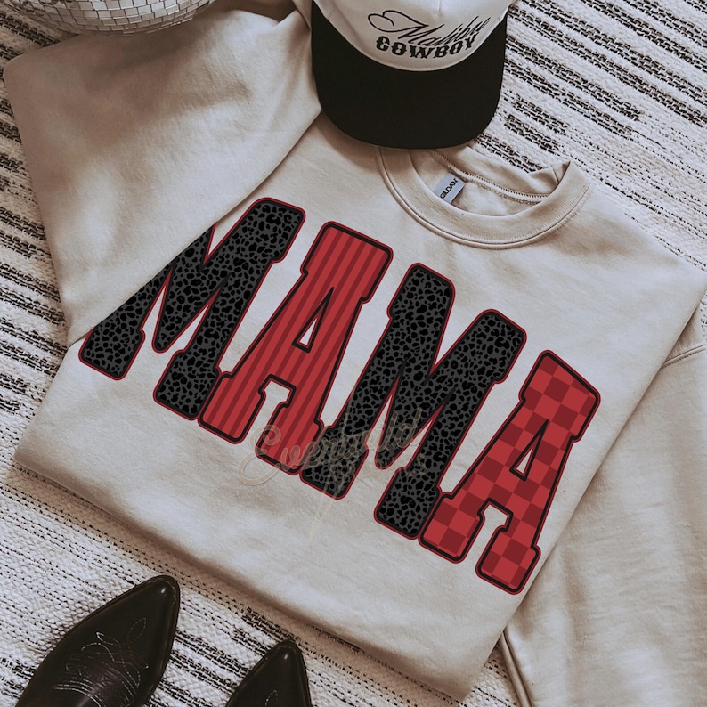 May include: A beige crewneck sweatshirt featuring the word MAMA in large block letters. The letters alternate between a black leopard print pattern and a red striped or checkered pattern, all outlined in a dark red border. A cap is placed above.