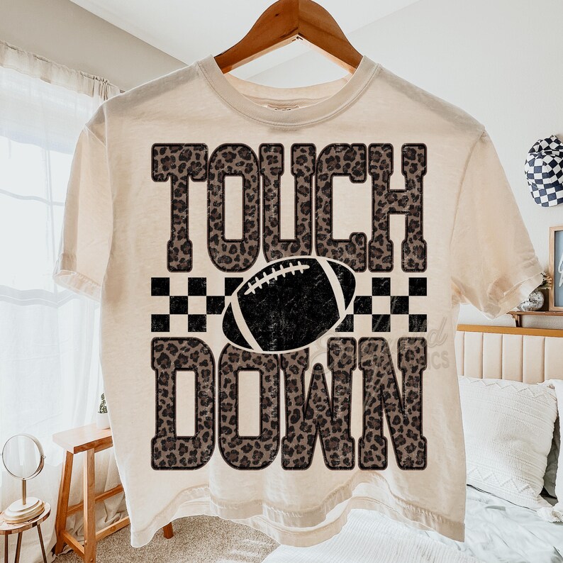 Puede incluir: Una camiseta de manga corta color crema con el texto TOUCH DOWN en un patr&oacute;n de leopardo marr&oacute;n. Entre las palabras hay un gr&aacute;fico de un bal&oacute;n de f&uacute;tbol americano en blanco y negro centrado sobre un patr&oacute;n de cuadros blanco y negro.