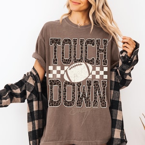 Puede incluir: Una camiseta marr&oacute;n de manga corta con el texto TOUCH DOWN en estampado de leopardo. Entre las palabras hay un gr&aacute;fico de un bal&oacute;n de f&uacute;tbol americano blanco centrado sobre un patr&oacute;n de cuadros blancos y marrones. Se lleva una camisa de cuadros negra y marr&oacute;n encima.