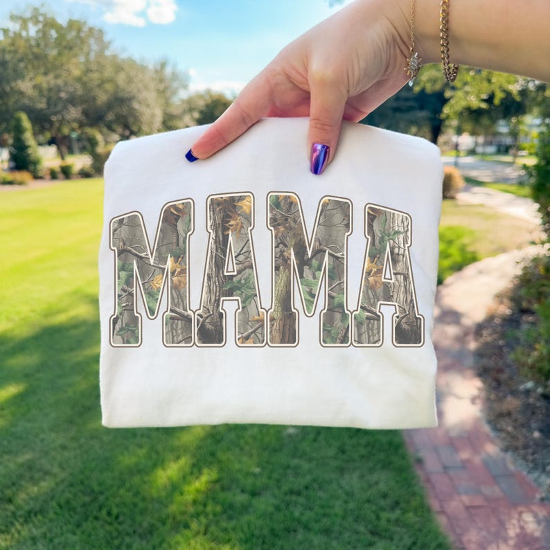 May include: A white sweatshirt with the word "MAMA" printed across the front in a camouflage pattern. The letters are in a bold, arched font. The sweatshirt is held up by a person.