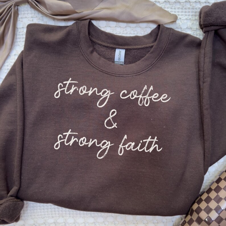 May include: A brown sweatshirt with the words "strong coffee & strong faith" in white script. The crew neck sweatshirt is made of a soft material and is suitable for casual wear.