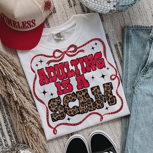 May include: White t-shirt with the slogan "ADULTING IS A SCAM" in red and leopard print lettering. The text is framed by a red and black ribbon design. A red and tan baseball cap with the word "TIMELESS" is also visible.