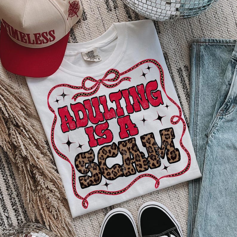 May include: White t-shirt with the slogan "ADULTING IS A SCAM" in red and leopard print lettering. The text is framed by a red and black ribbon design. A red and tan baseball cap with the word "TIMELESS" is also visible.
