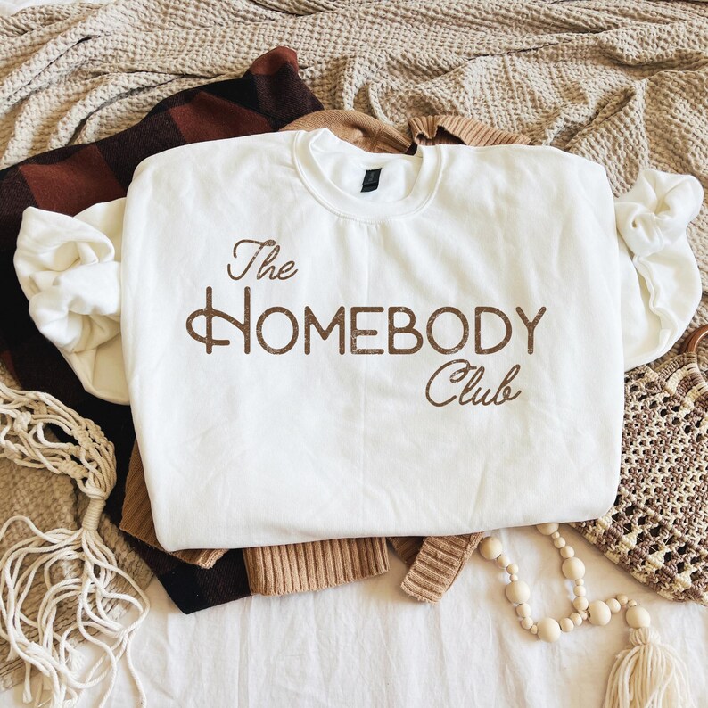 K&ouml;nnte beinhalten: Ein wei&szlig;es Sweatshirt mit Rundhalsausschnitt und dem braunen Text "The Homebody Club". Das Sweatshirt liegt gefaltet auf anderen Kleidungsst&uuml;cken, darunter eine braun-schwarz karierte Decke und ein beigefarbener Pullover. Eine Makramee-Tasche und eine Perlenkette sind ebenfalls zu sehen.