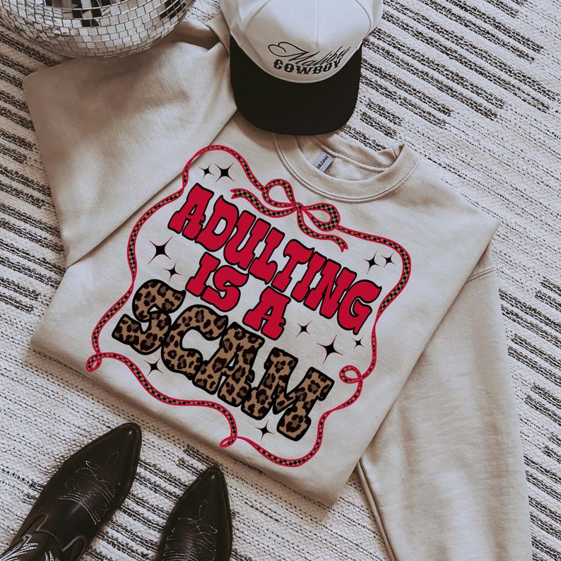 May include: A beige sweatshirt with the phrase "ADULTING IS A SCAM" in red and leopard print. A white and black trucker hat and black cowboy boots are also in the image.