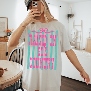 May include: Oversized beige t-shirt with the text "RAISED ON 90'S COUNTRY" in bright pink, set against a backdrop of teal vertical stripes. The shirt has a relaxed fit and short sleeves. A black baseball cap is worn.
