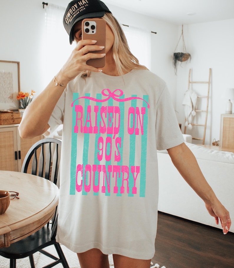 May include: Oversized beige t-shirt with the text "RAISED ON 90'S COUNTRY" in bright pink, set against a backdrop of teal vertical stripes. The shirt has a relaxed fit and short sleeves. A black baseball cap is worn.