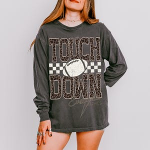 Puede incluir: Una camiseta de manga larga gris carb&oacute;n con el texto TOUCH DOWN en un patr&oacute;n de estampado de leopardo marr&oacute;n. Entre las palabras hay un gr&aacute;fico de bal&oacute;n de f&uacute;tbol americano blanco sobre un fondo de cuadros negros y blancos.